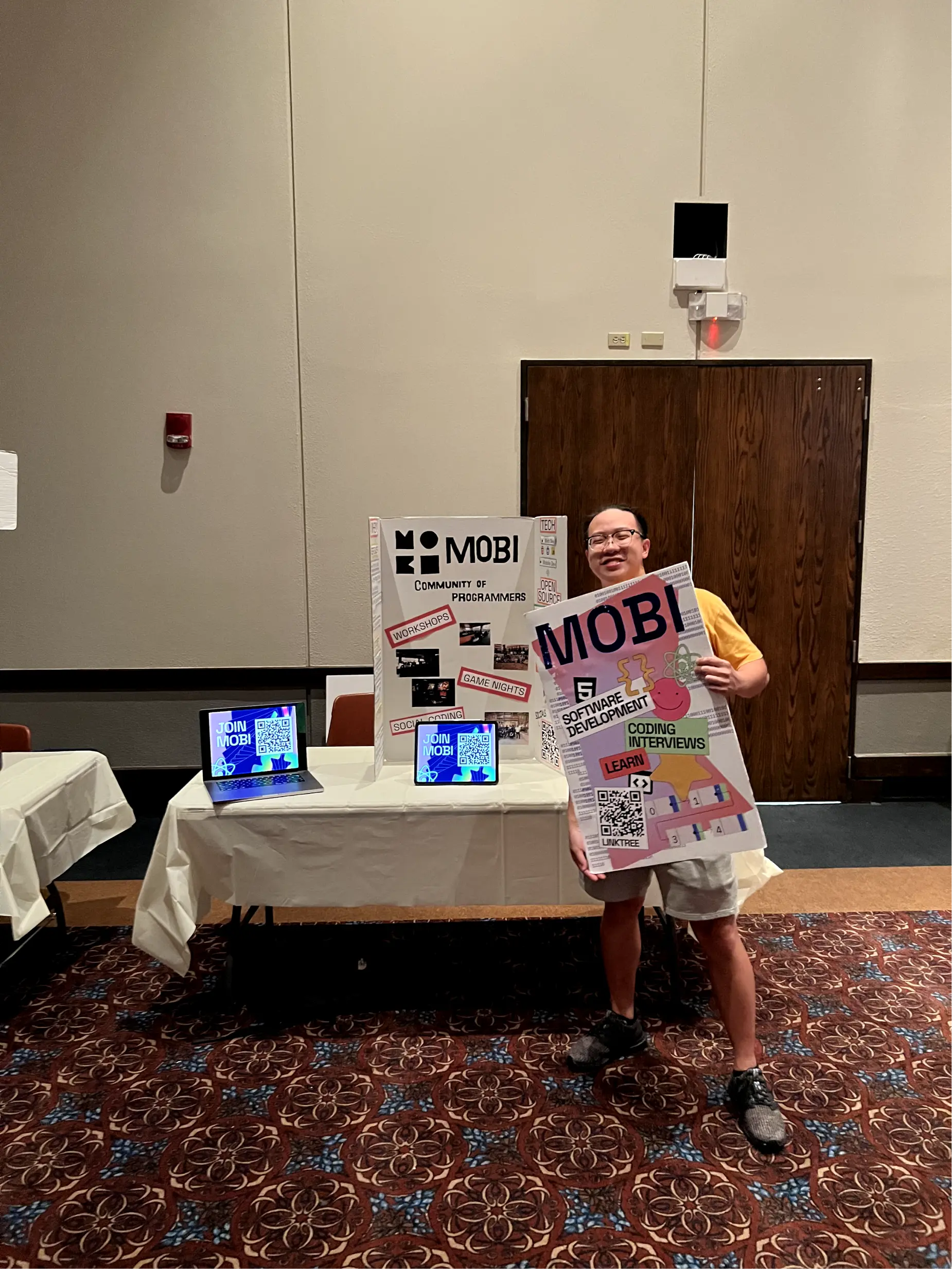 Long Nguyen standing and holding a large MOBI poster in front of a MOBI display. The display contains a laptop, iPad, and tri-board display of different graphics and information about MOBI.